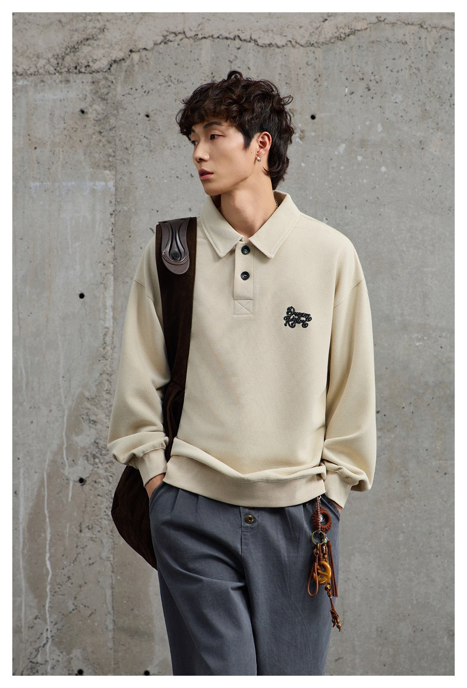 Model wearing a beige polo-collar sweatshirt with gray trousers against a concrete wall, carrying a brown bag