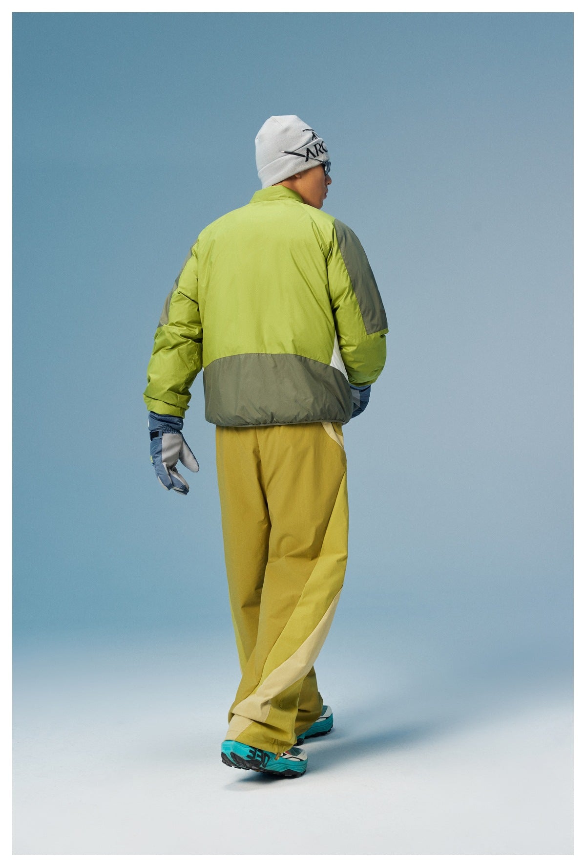 Rear view of a lime green jacket with gray shoulder and hem panels and a white side insert, worn with mustard trousers.