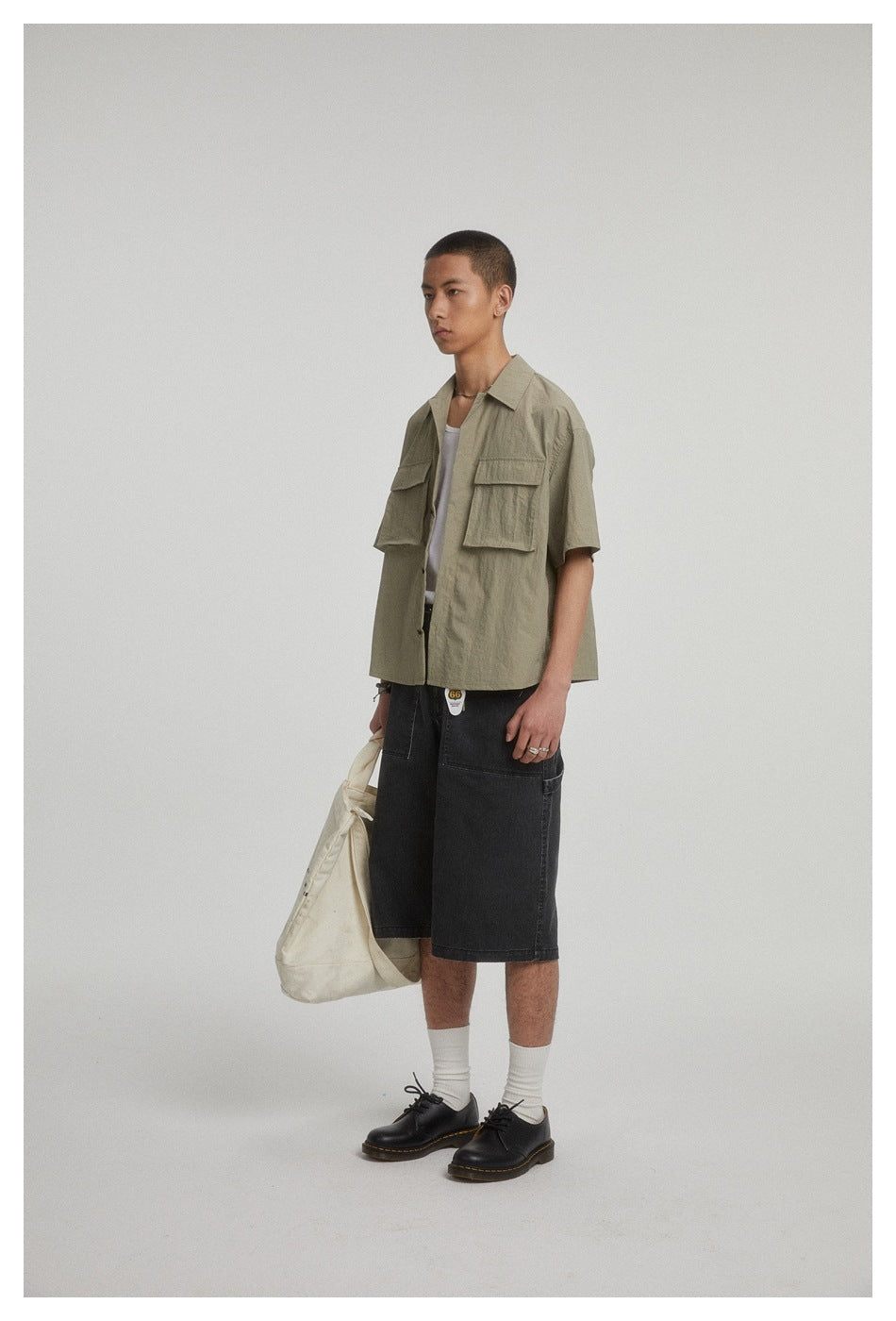 Model in black denim knee-length shorts with an olive short-sleeve shirt, holding a cream tote bag, side view