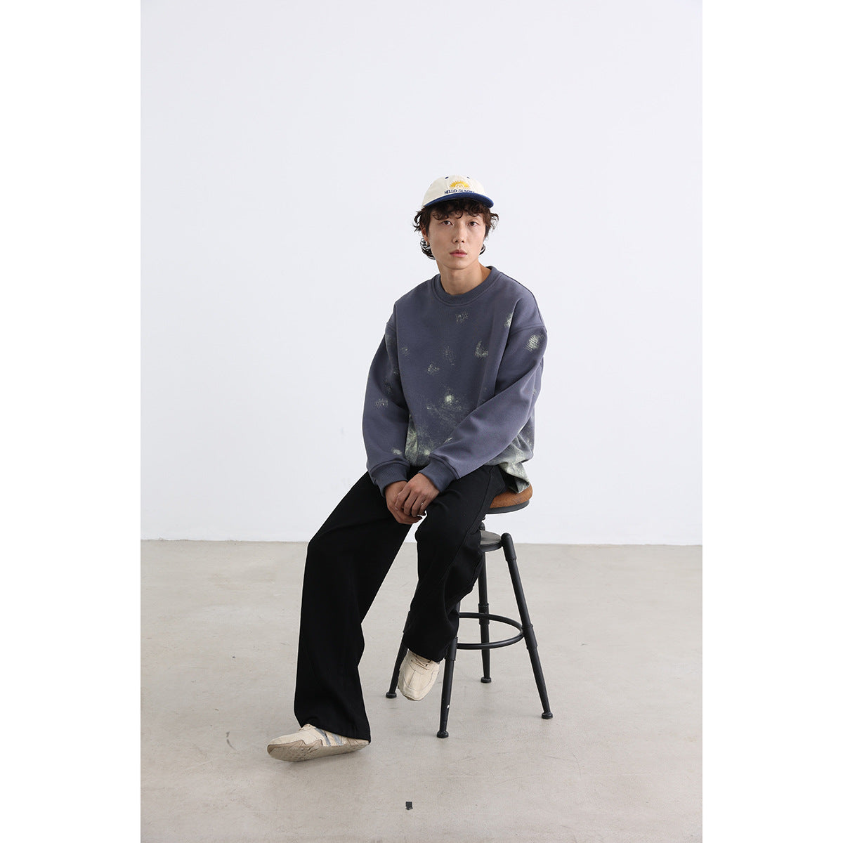 Model seated wearing an oversized slate-blue crewneck sweatshirt with scattered pale distressed graphics and black trousers.