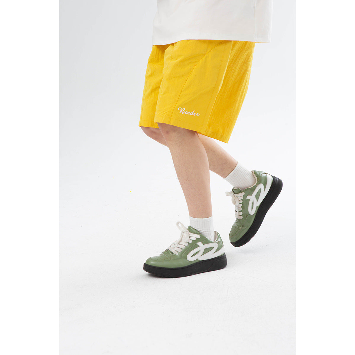 Close-up of bright yellow shorts worn with white socks and green sneakers; white script appears near the hem.
