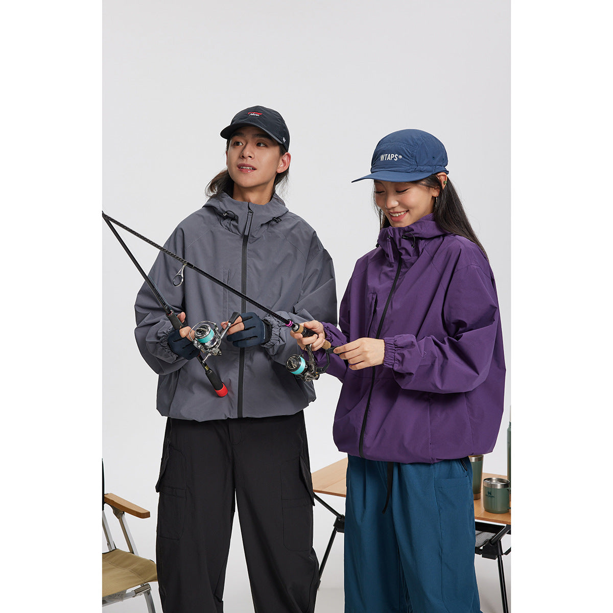 Two people wearing gray and purple hooded jackets hold fishing rods against a plain light background.