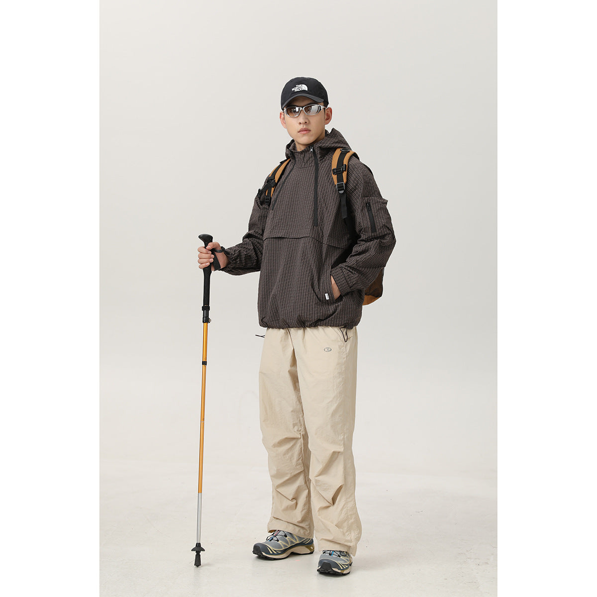 Full-length model wearing a dark brown checked hooded pullover jacket and beige trousers, with a backpack and trekking pole.
