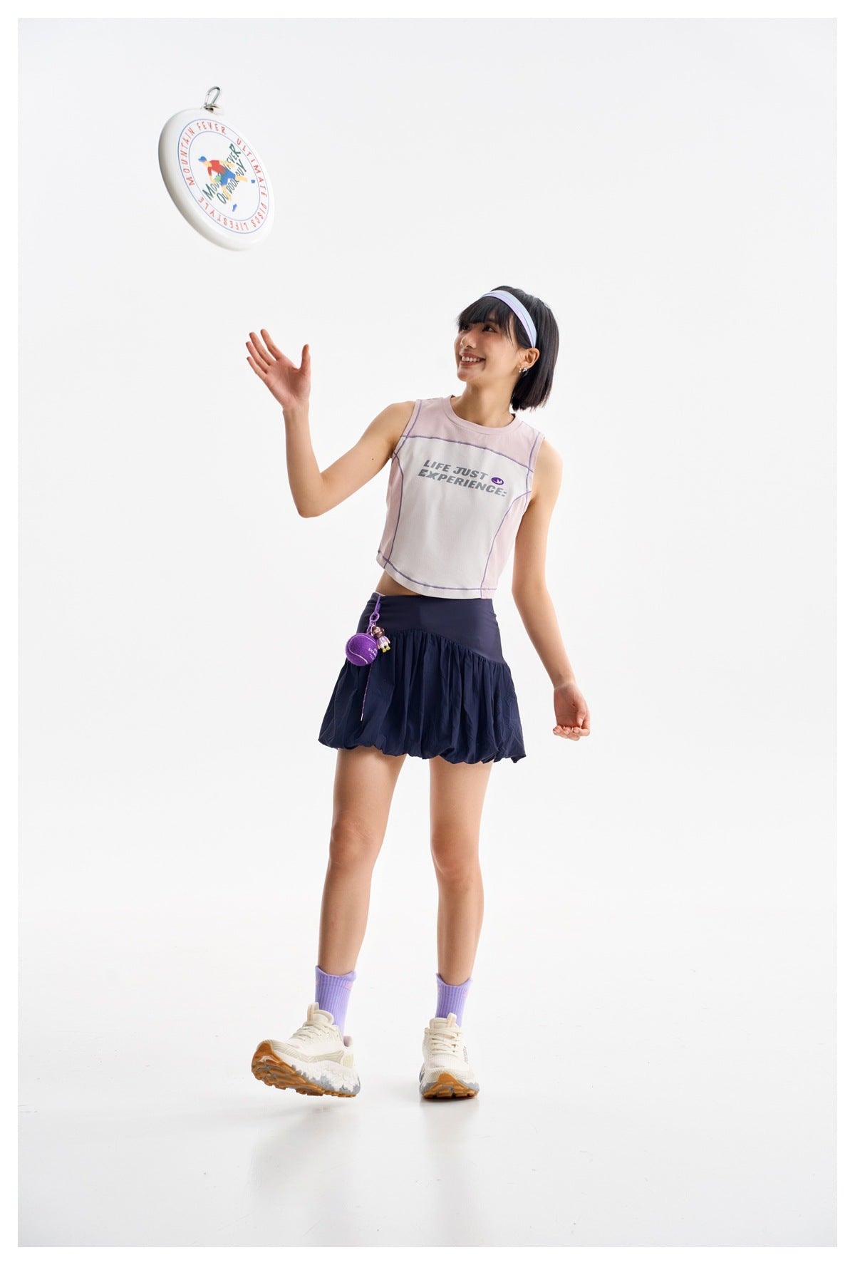 A model in a pale pink sleeveless crop top and navy bubble skirt reaches toward a flying disc.
