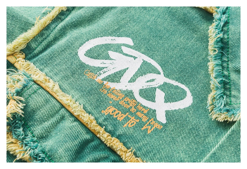 Close-up of teal denim with yellow-and-teal frayed seams and a large white graphic with small orange lettering.