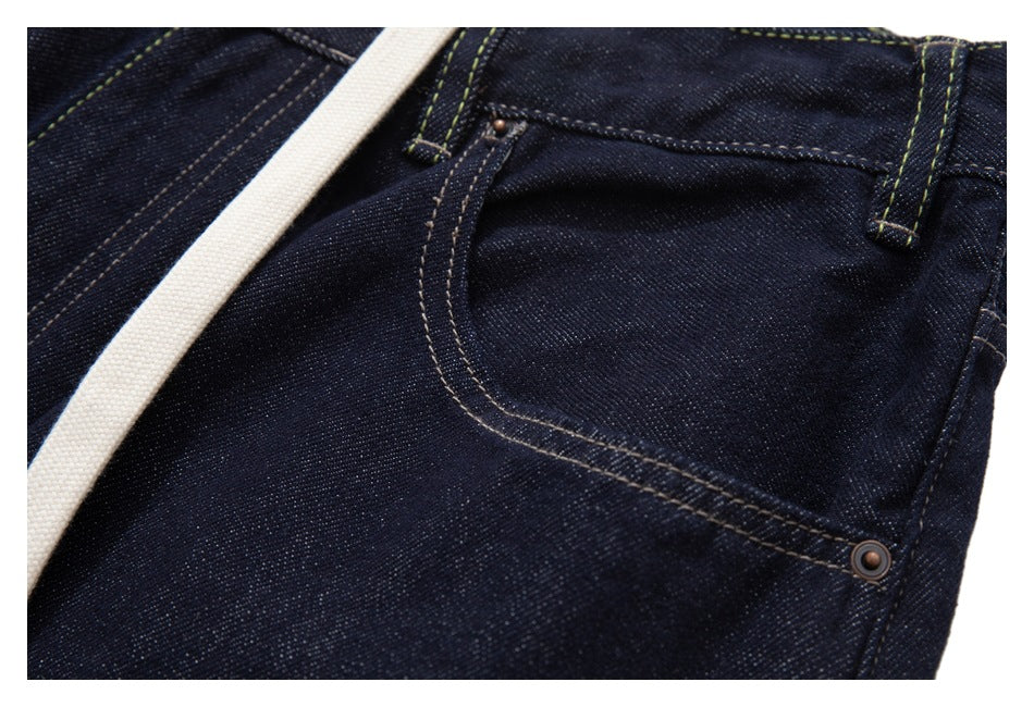 Close-up of a riveted front pocket on dark indigo jeans, with contrast stitching and a white drawstring.