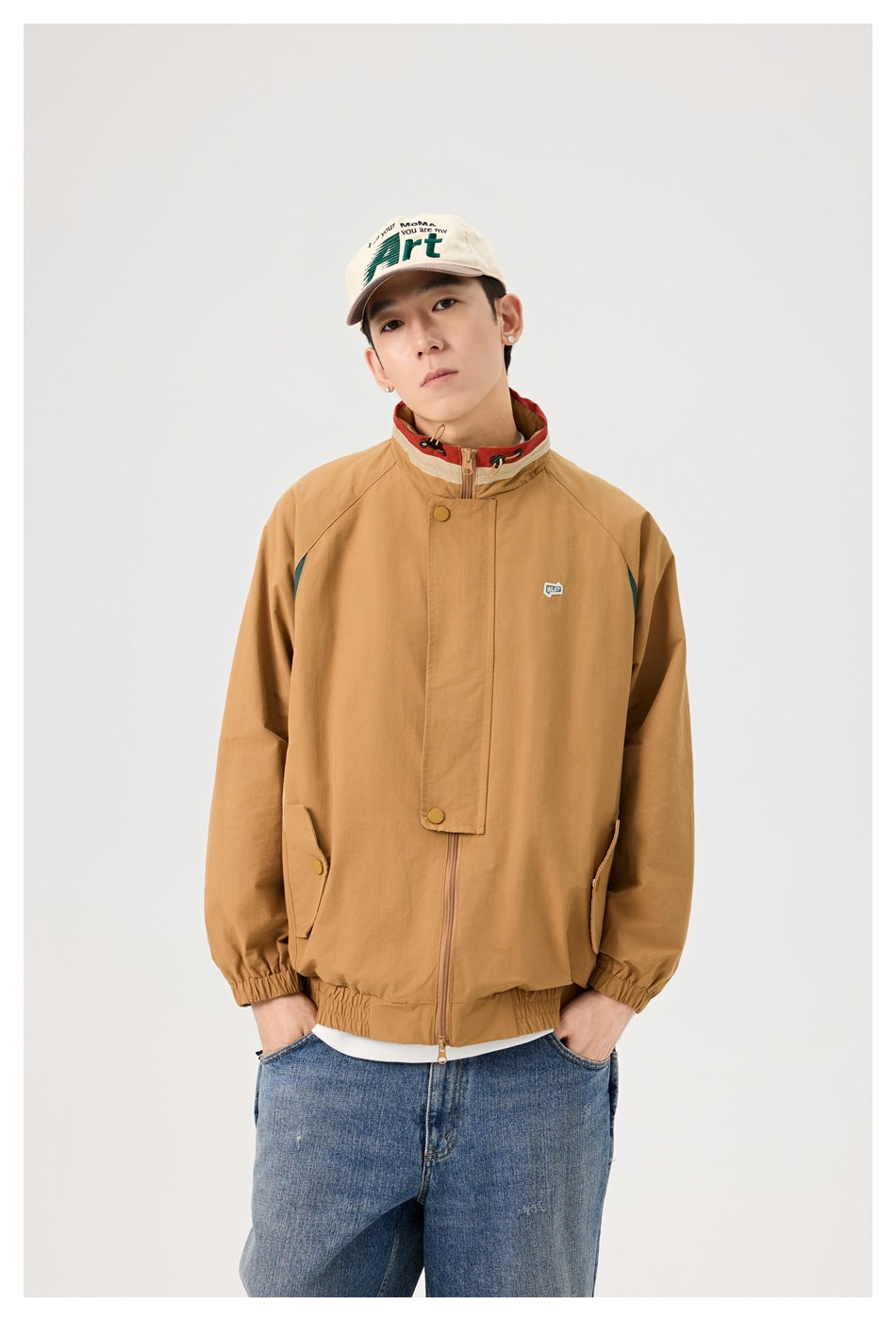 Model wearing a tan hooded jacket with a stand collar, snap-front storm flap, and dark teal side panels.