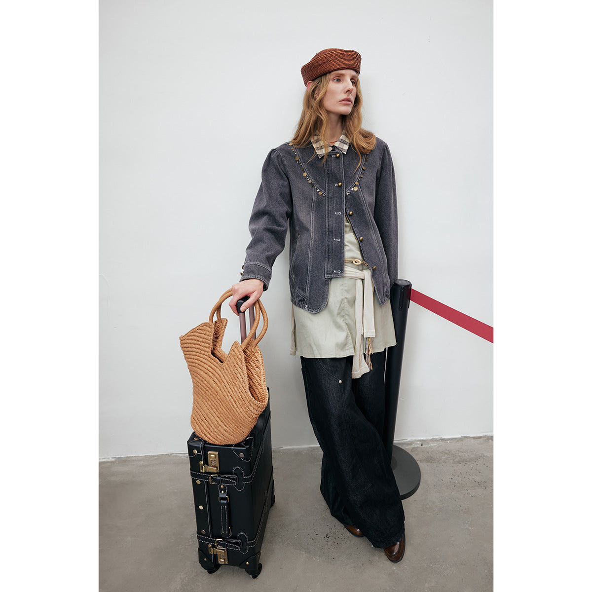 Model wearing a charcoal embellished denim jacket and wide-leg black trousers beside a black suitcase and woven tote.