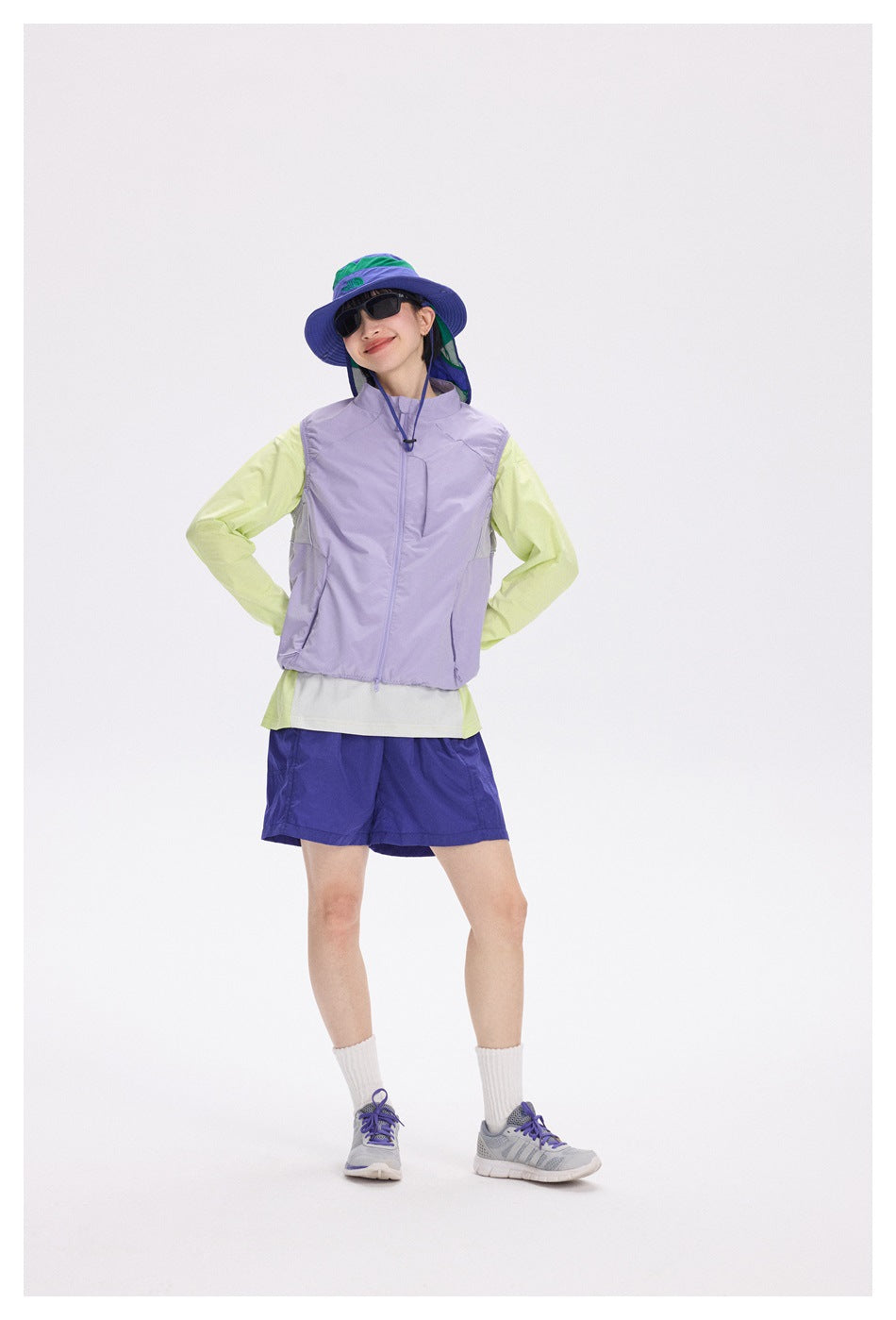 A model wears a lavender zip vest over a lime long-sleeve top, with blue shorts and a color-block sun hat.
