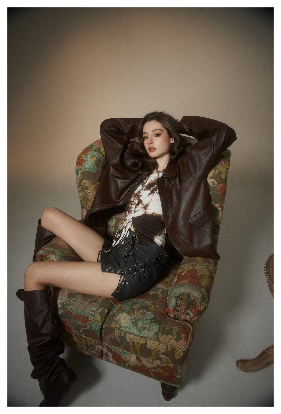 Seated model in an oversized dark brown jacket, printed top, black lace-up mini skirt, and tall boots.