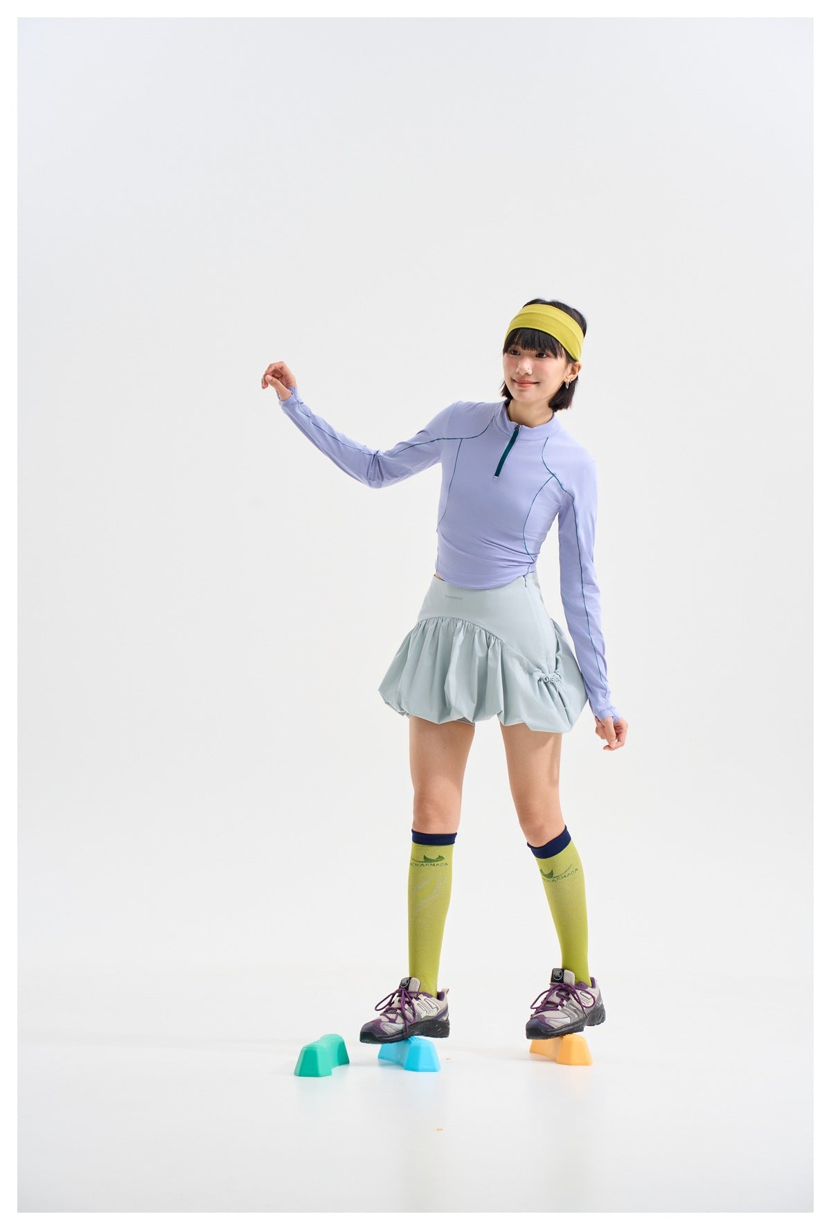 Model balancing on colorful step blocks in a lavender quarter-zip top and pale blue-gray gathered skirt.