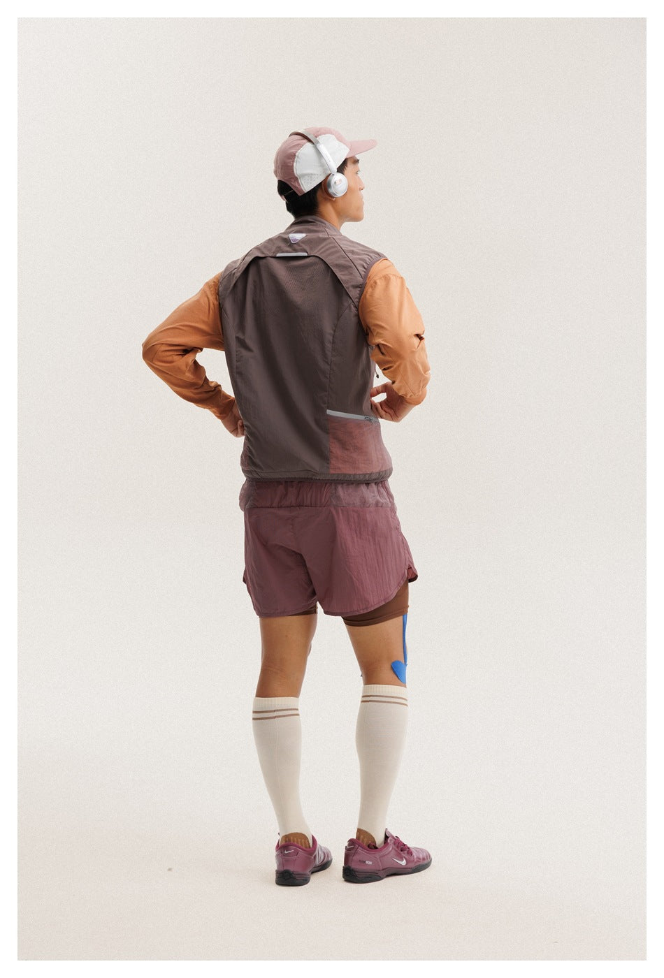 Rear view of a man wearing a dark taupe sleeveless vest over an orange top and mauve shorts.