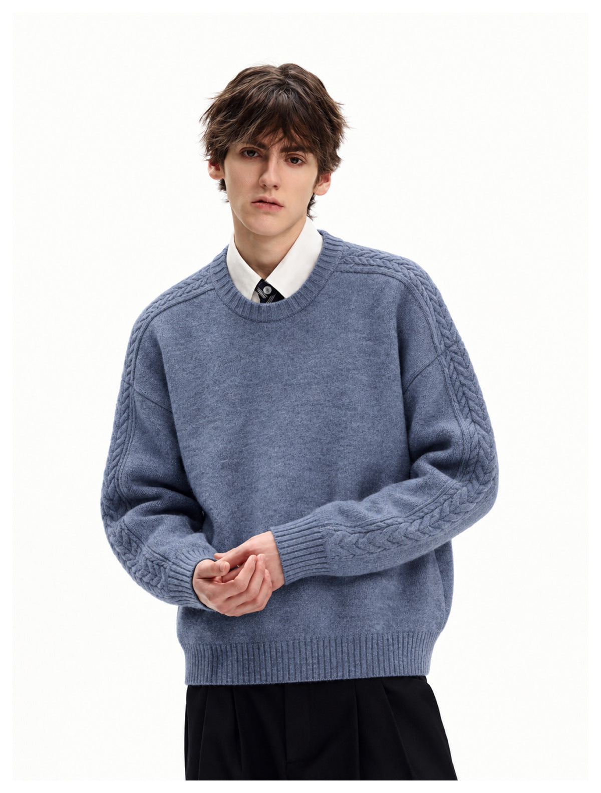 Model wearing blue cable-knit crew-neck sweater over a white collared shirt and tie, hands clasped