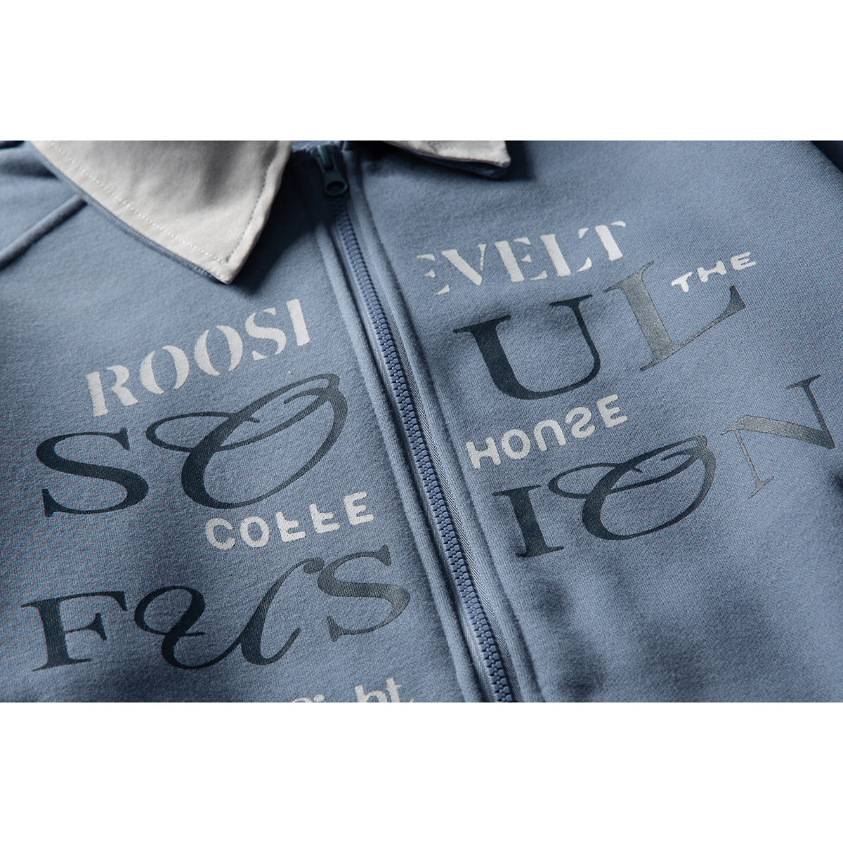 Close-up of a blue zip-up jacket showing its zipper, light gray collar, and large multicolored lettering.