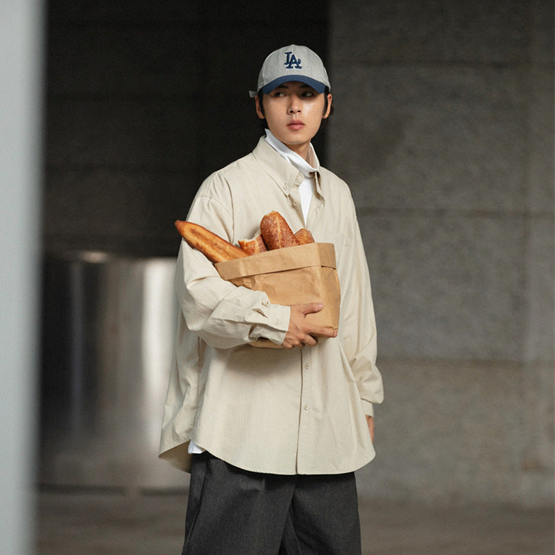 Model wearing a loose cream button-up shirt and dark trousers, holding a paper bag of bread.