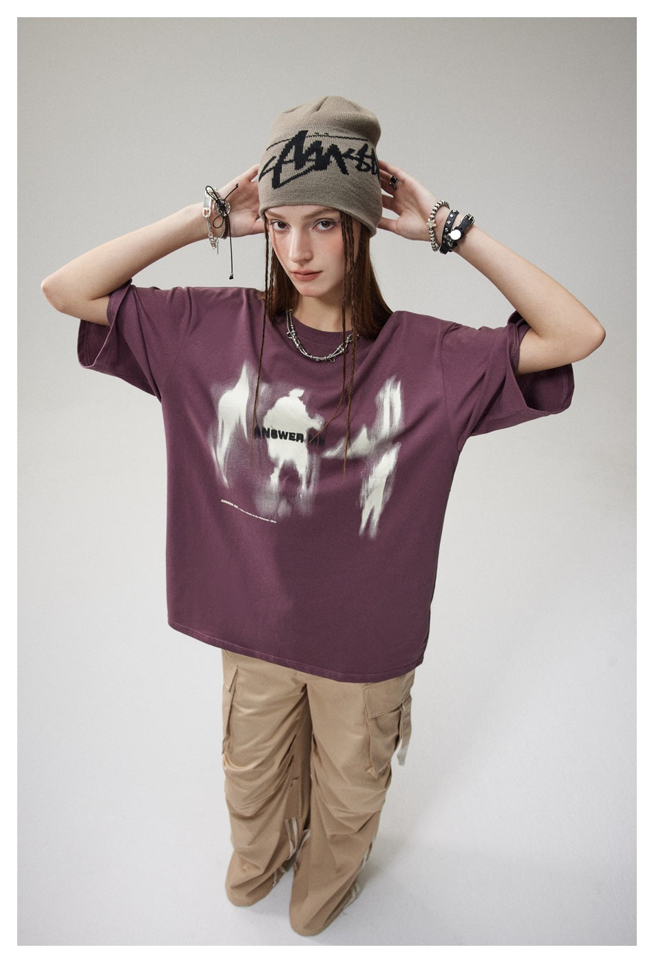 Full-length model wearing an oversized plum graphic T-shirt, taupe beanie, and loose tan cargo pants.