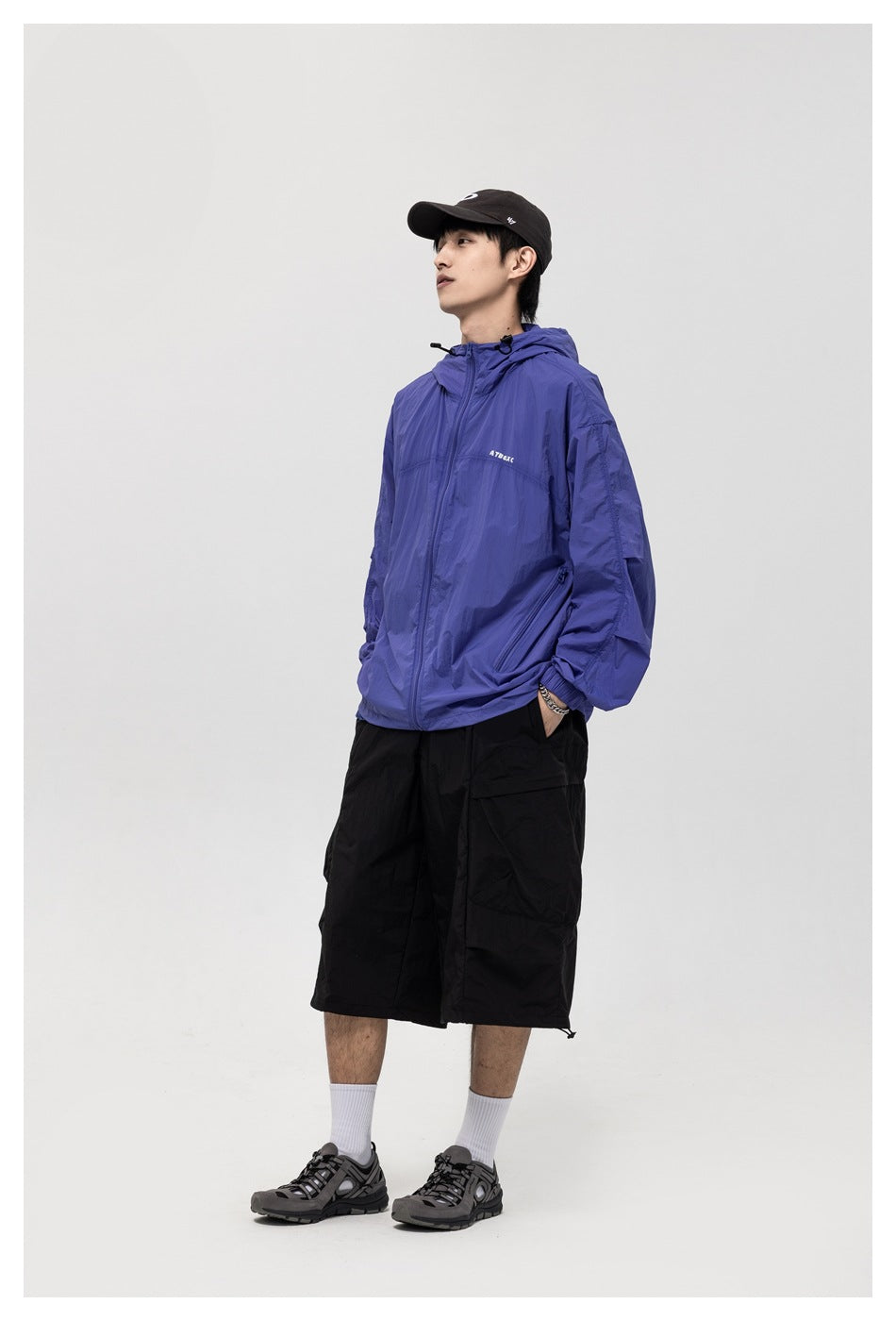 Model wearing a blue hooded zip jacket with black cargo shorts, cap and sneakers, angled side view