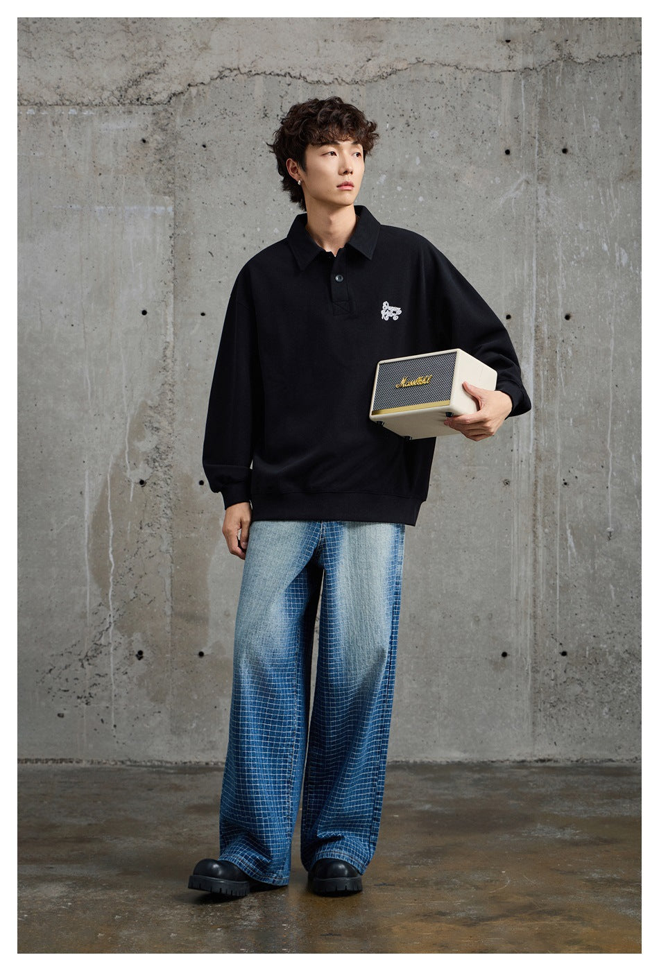 Full-length model wearing a black oversized collared pullover with white chest embroidery and wide blue checked jeans.