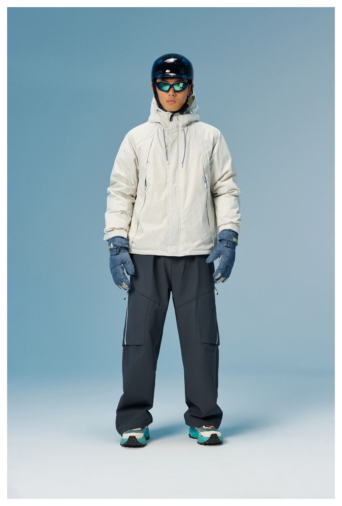 Model wearing a white subtly striped hooded ski jacket, dark snow pants, blue gloves, helmet and goggles, front view.
