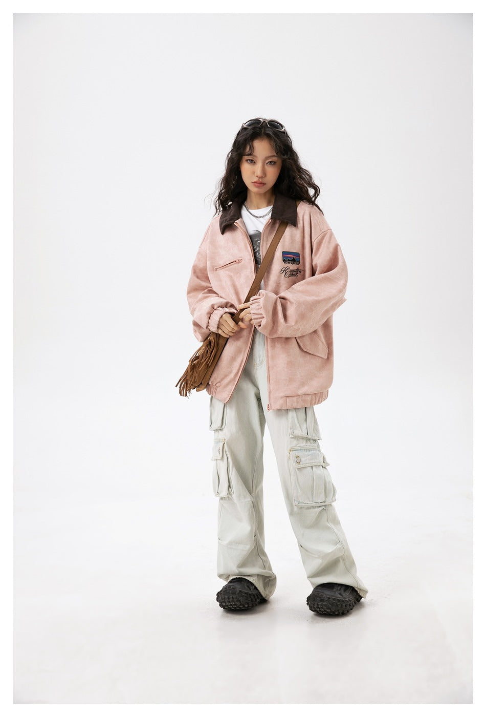 Full-length front view of a model in a dusty pink jacket and pale cargo pants, carrying a brown fringed bag.