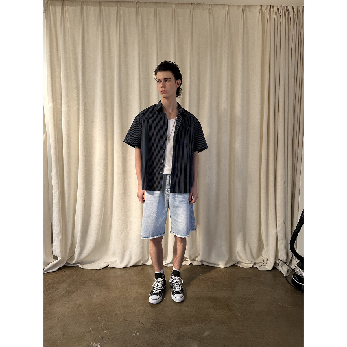 Model wearing a dark pinstriped short-sleeve button-up shirt over a white tank and light blue frayed denim shorts, front