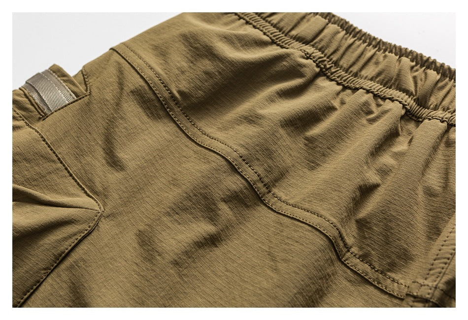 Close-up of olive-brown trousers showing the elastic waistband, stitched pocket, and narrow webbing loop.