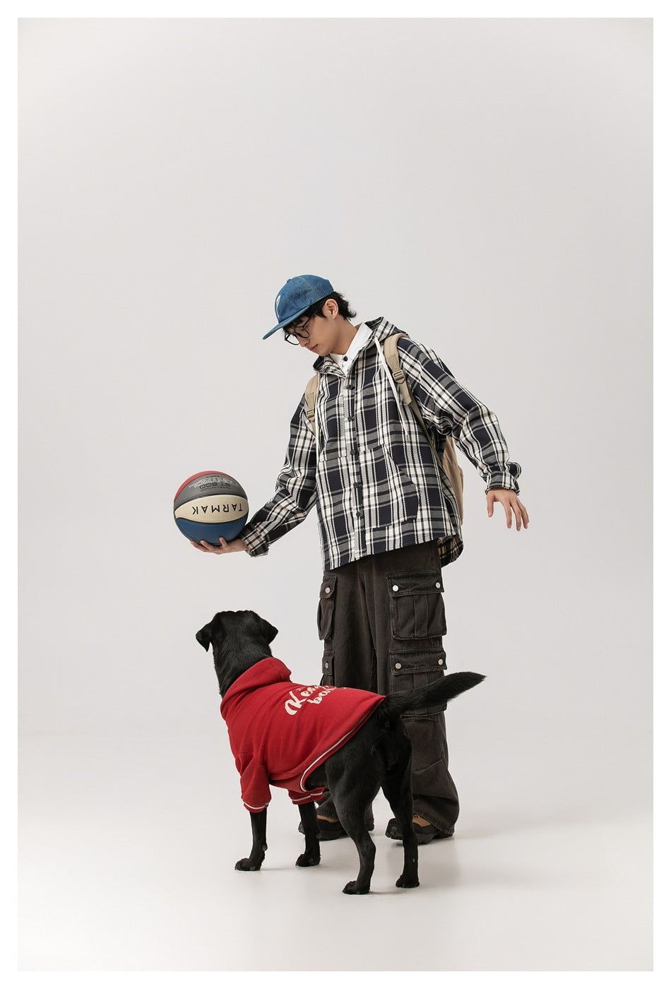 Model wearing a black-and-white plaid hooded jacket and dark cargo pants, holding a basketball beside a dog.