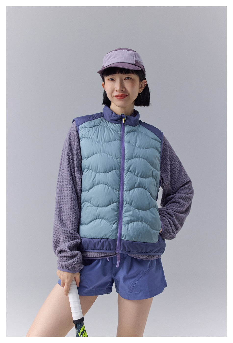 Closer front view of a model wearing a pale blue quilted vest with a purple zipper and trim over a textured purple top.