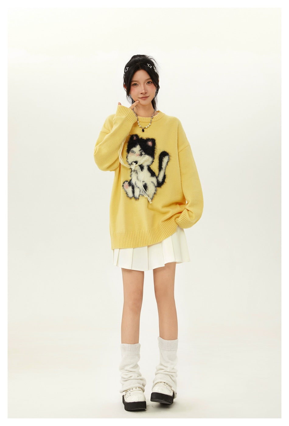 Model wearing an oversized yellow crewneck sweater with a fuzzy black-and-white cat graphic and white pleated skirt.