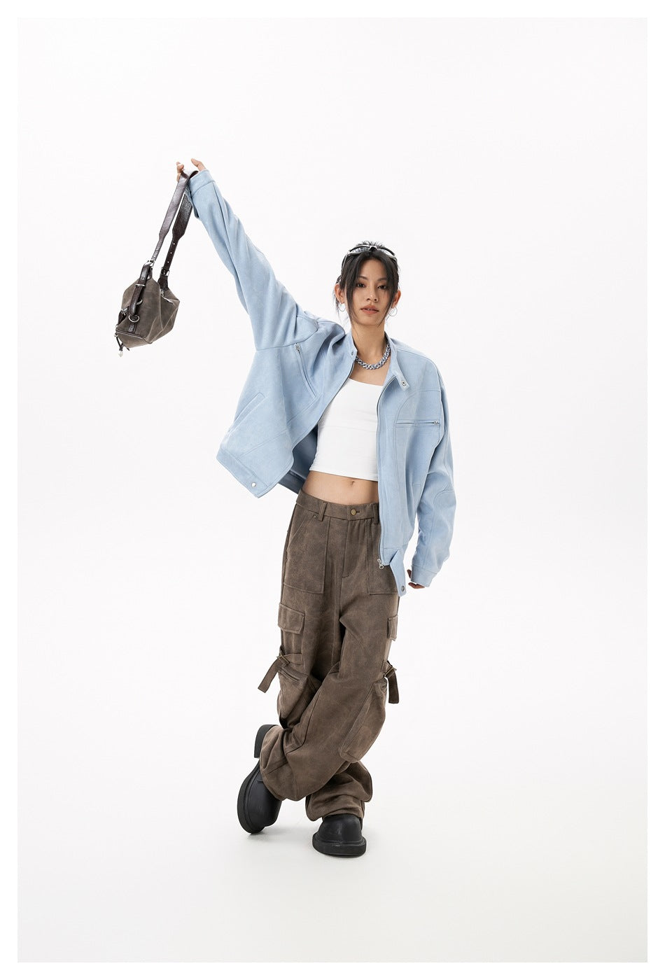 Model wearing a light blue jacket and brown cargo pants, posing with one arm raised while holding a small bag.