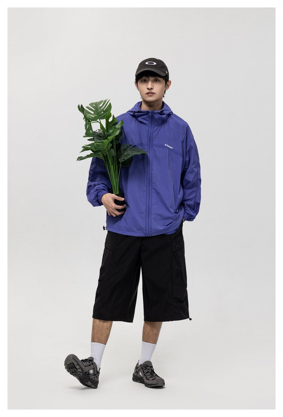 Full-length view of a model wearing a blue-violet hooded zip jacket and black shorts, holding a leafy plant.