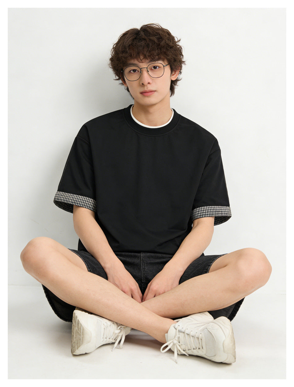Seated model wearing a black tee with cream neckline trim and plaid cuffs with dark denim shorts