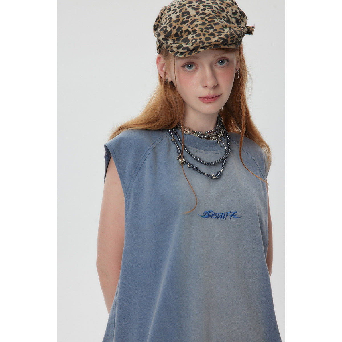Model wearing a slate-blue sleeveless top with small blue chest embroidery, layered necklaces and a leopard-print cap.