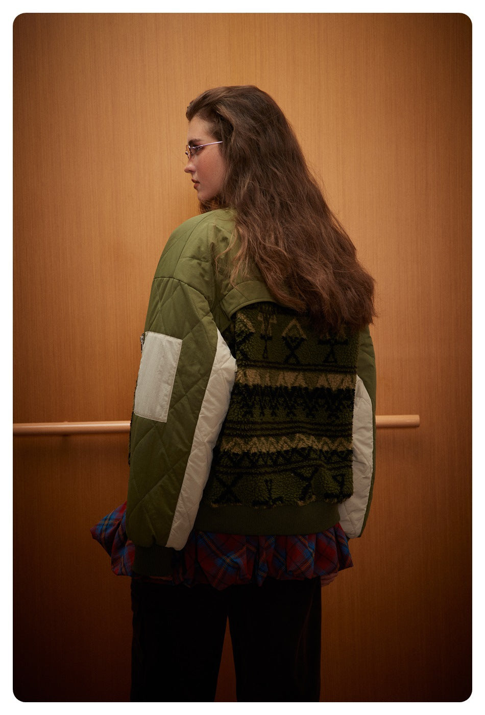 Rear view of a model wearing an olive quilted bomber jacket with cream sleeve panels and a dark patterned fleece back.