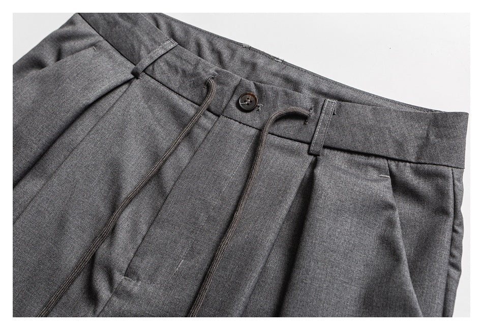Close view of charcoal gray trousers with a buttoned waistband, belt loops, front pleats, and a drawstring.
