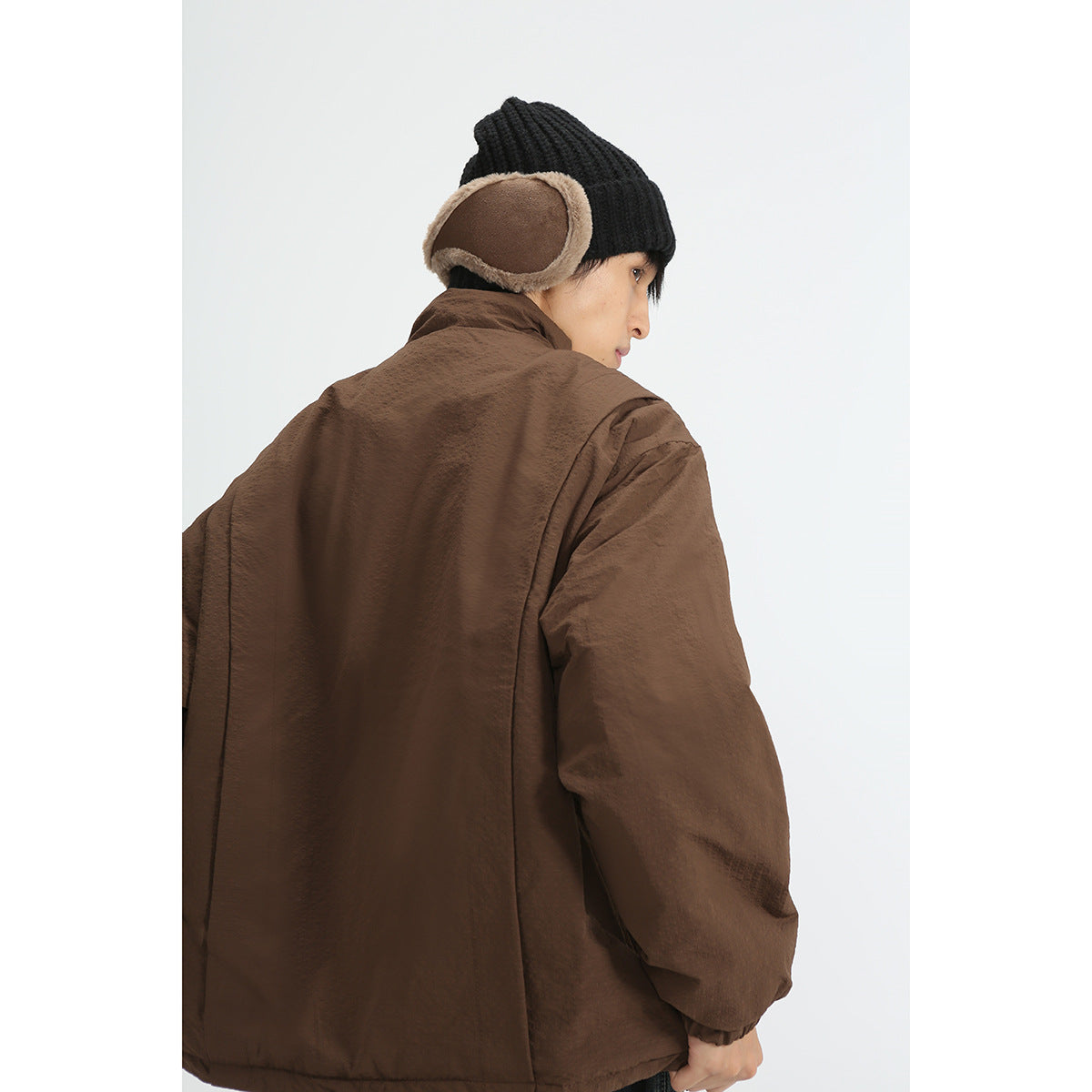 Rear view of a model wearing a brown jacket with a high collar and loose sleeves, paired with a black knit earflap hat.