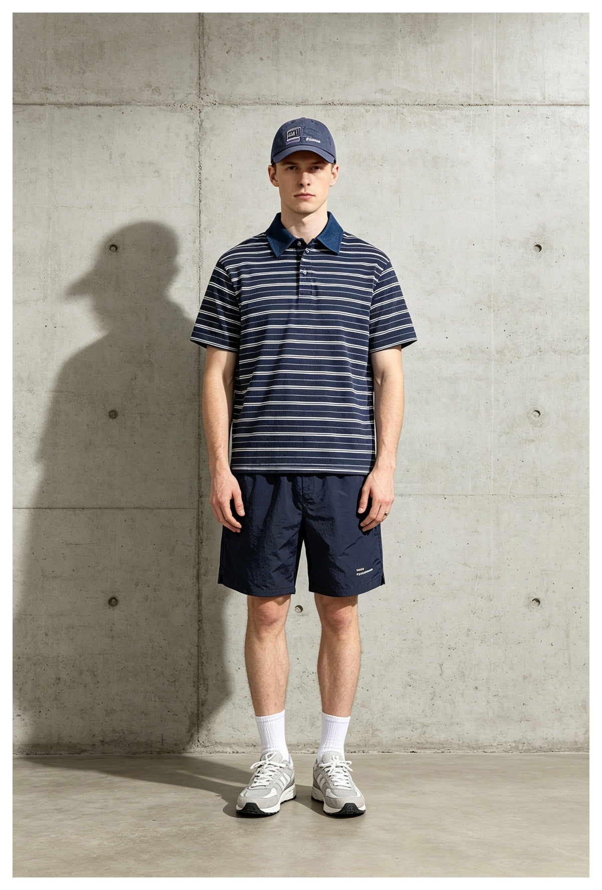 Man standing against a concrete wall in a navy striped polo shirt with blue collar, navy shorts and a cap
