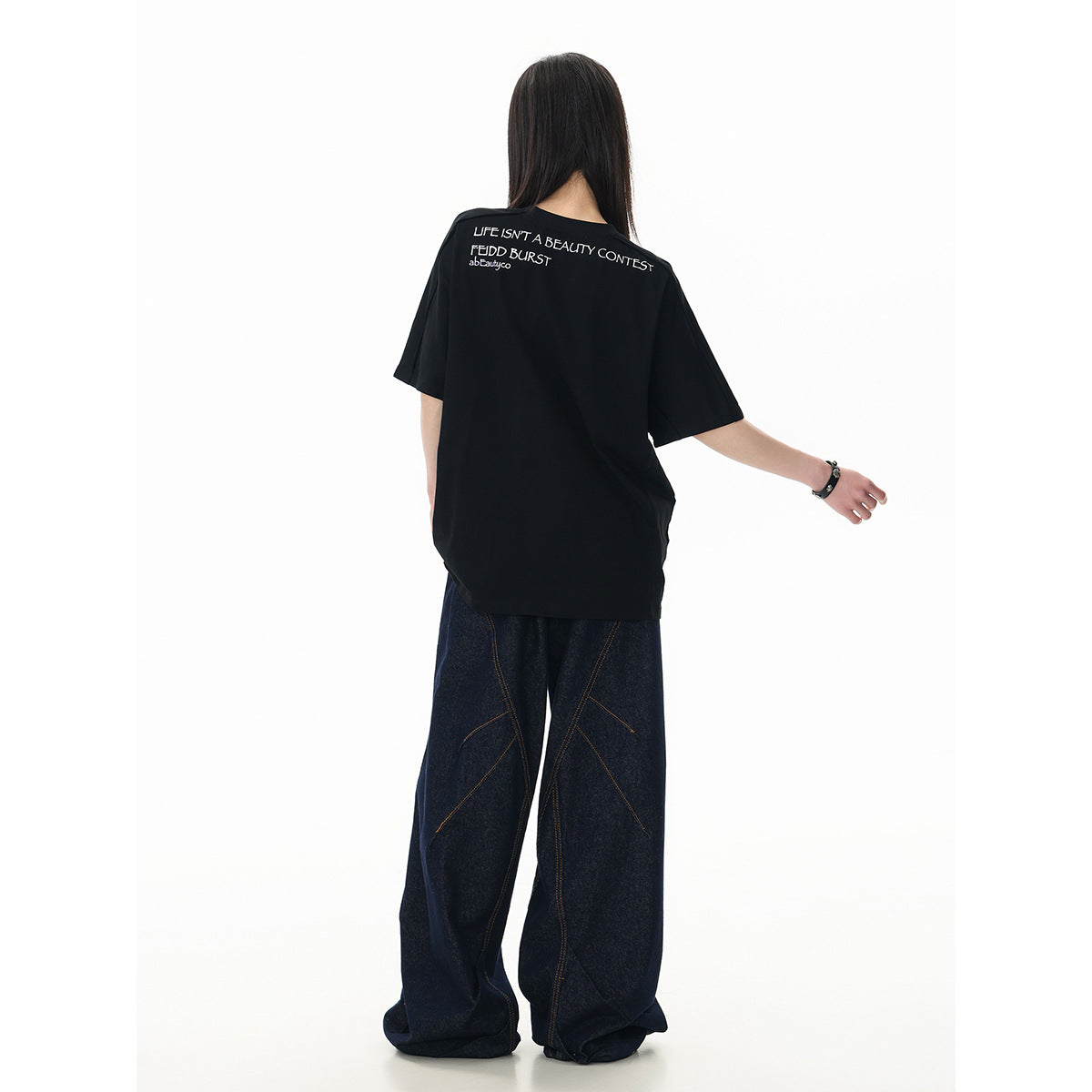 Rear view of a model wearing a black oversized T-shirt with white lettering across the upper back and dark wide-leg jeans.