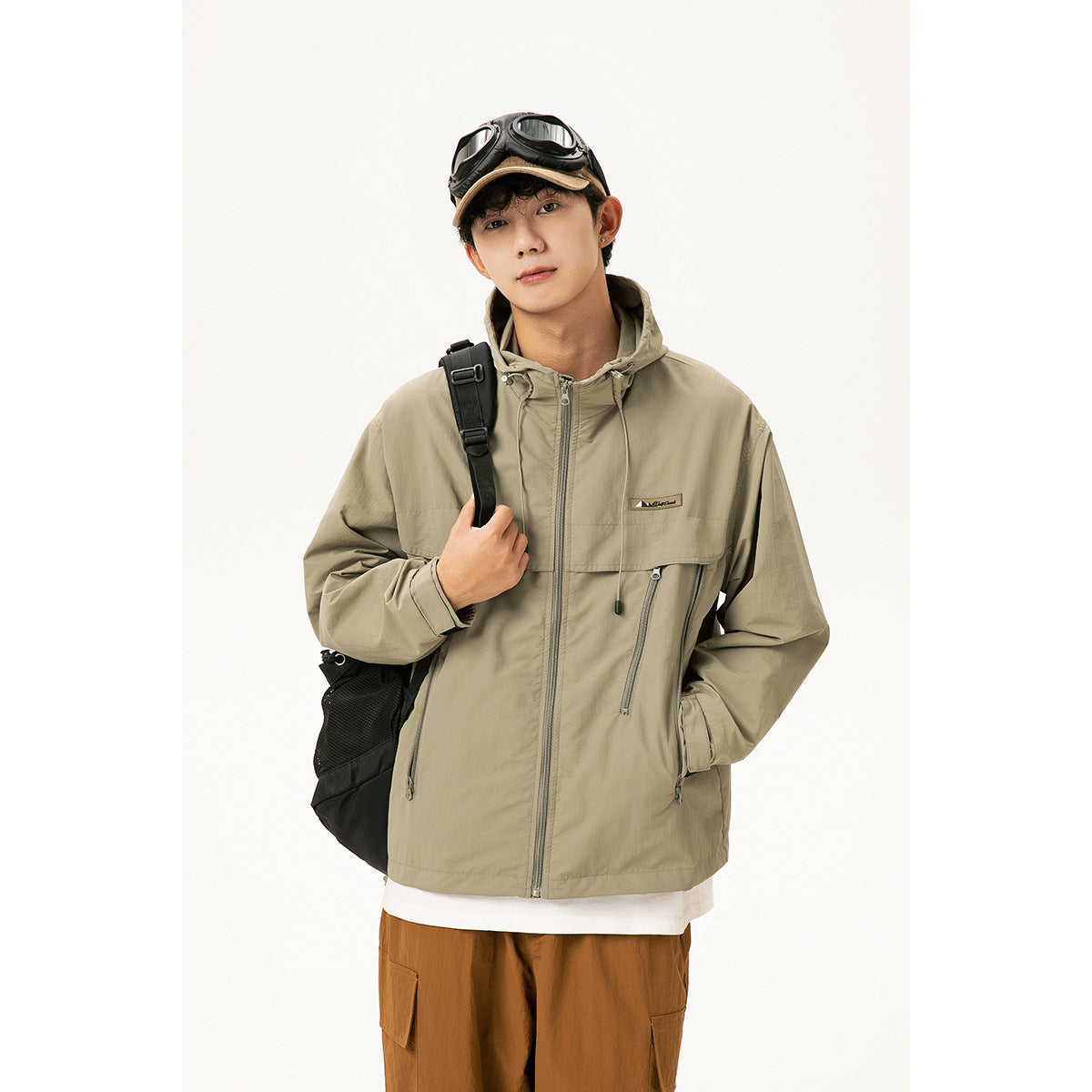 Model wearing a sage-gray hooded zip jacket with multiple pockets, brown cargo pants, and a cap with goggles.