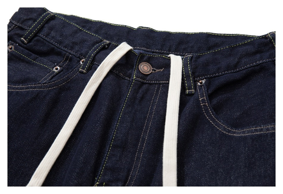 Close-up of dark indigo jeans with a button waist, belt loops, riveted pockets, contrast stitching and white drawstrings.
