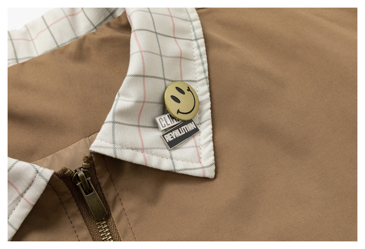 Close-up of a smiley pin and text badges attached to the pale plaid collar of the tan jacket.