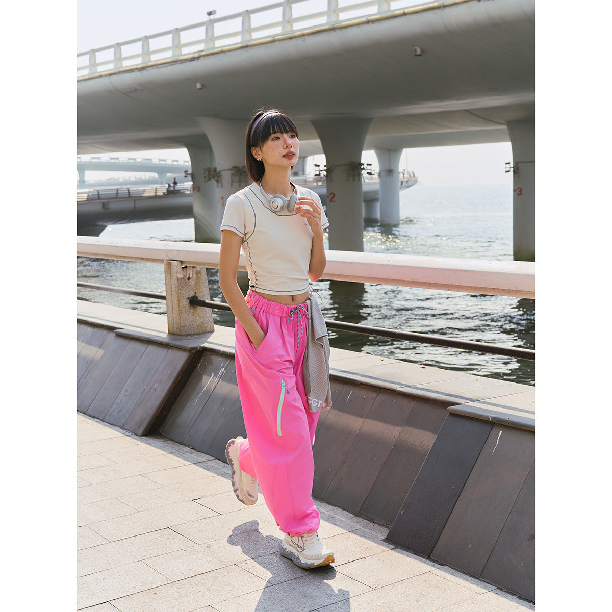 Model wearing bright pink loose trousers with a cream fitted top beside a waterfront.