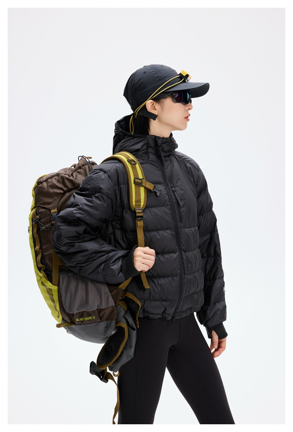 Closer view of a model wearing a black hooded quilted jacket, shown with a hiking backpack and cap.