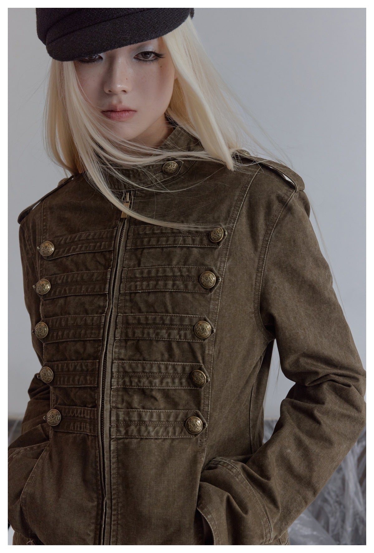 Close view of a model wearing a dark olive-brown jacket with a high collar, zipper, and decorative button tabs.