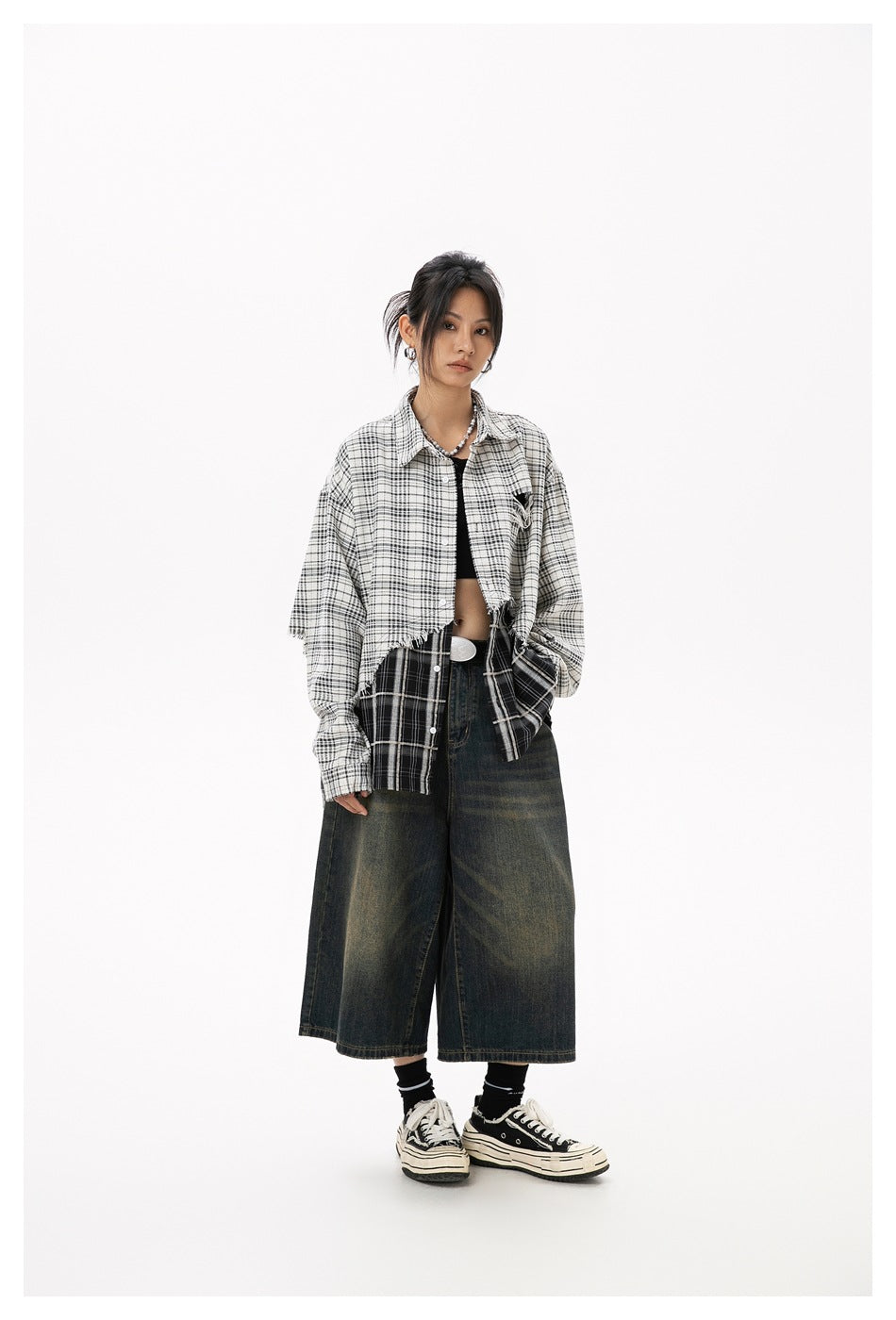 Full-length front view of a model in an oversized layered plaid shirt with a frayed hem and dark wide-leg cropped jeans.
