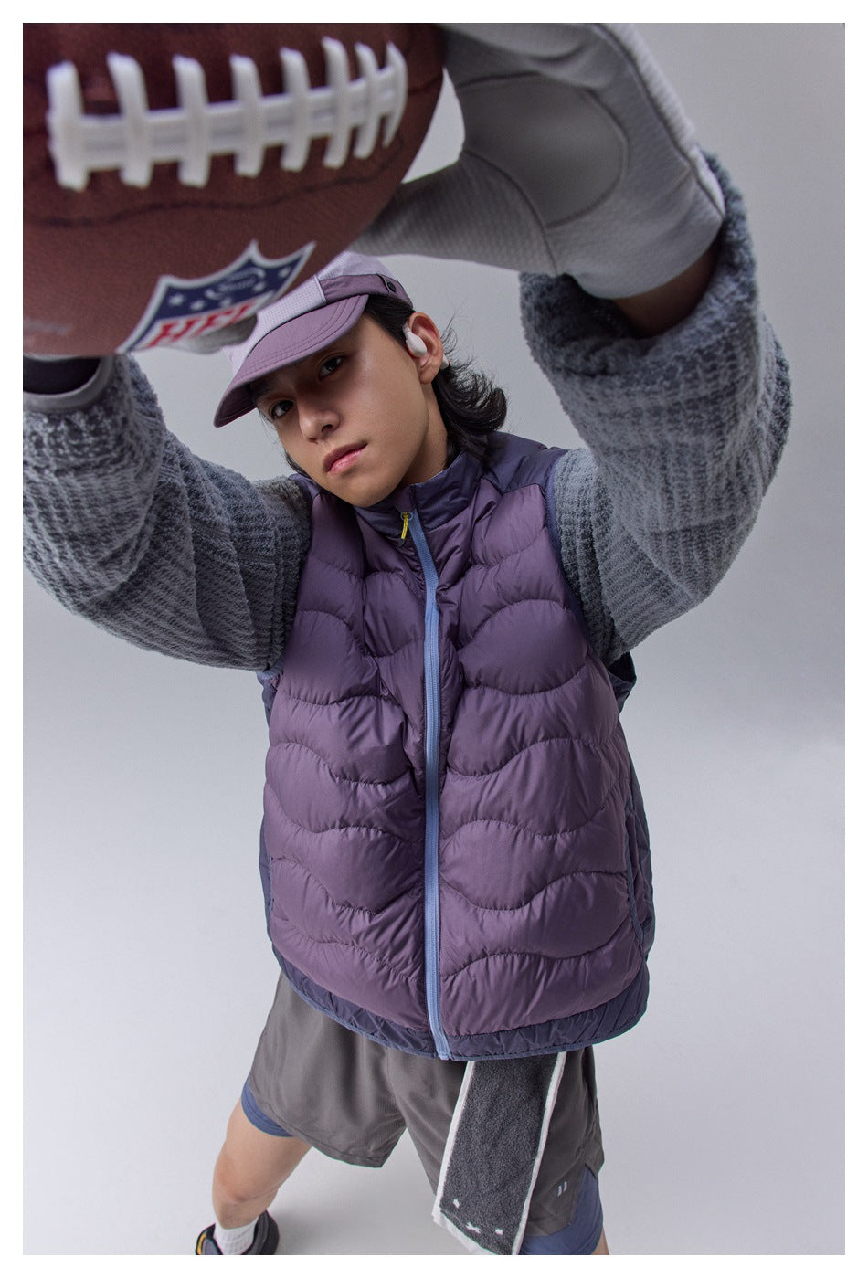 Close front view of a model wearing a purple quilted vest and gray pullover while reaching for a football.