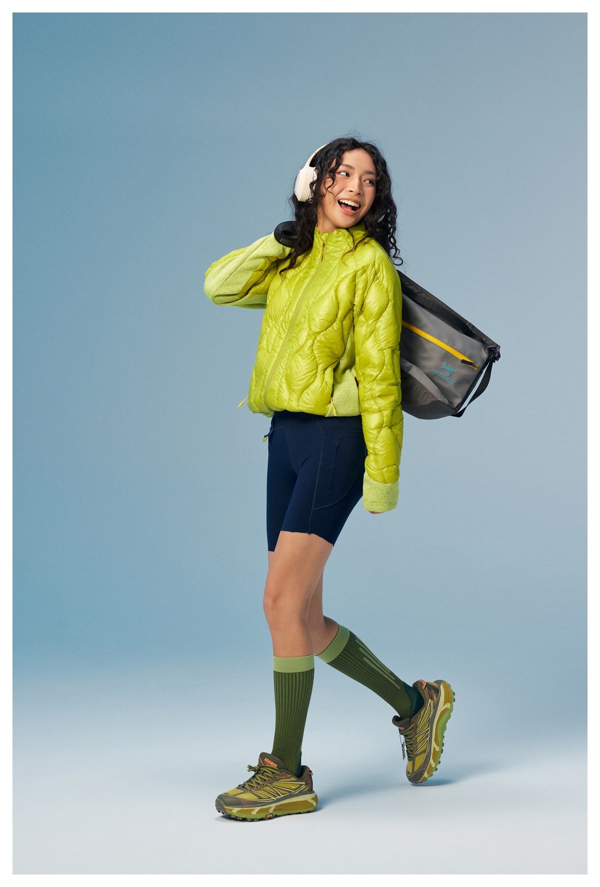 Model in a yellow-green quilted zip jacket with navy shorts, headphones and a gray shoulder bag, looking back