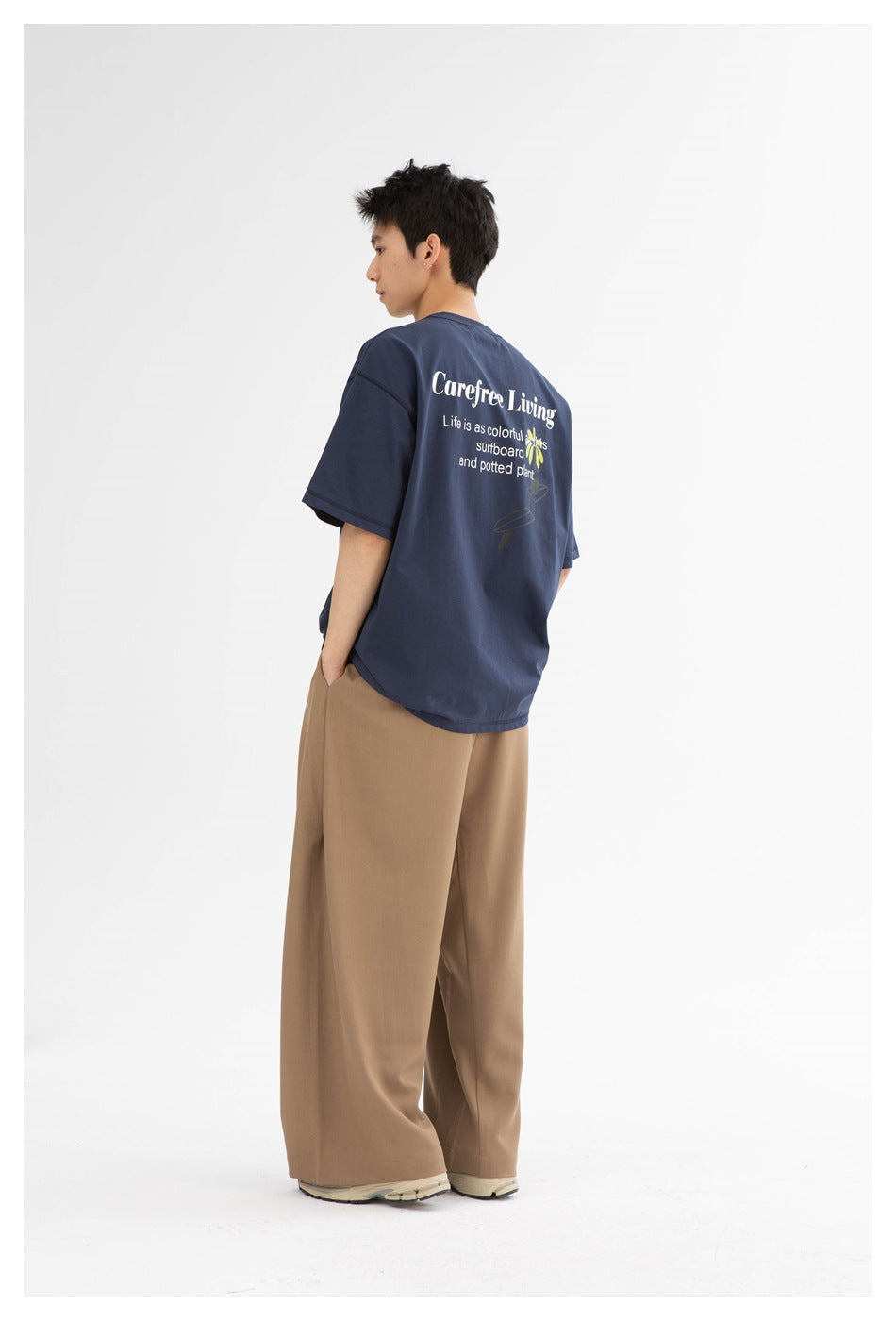 Model in a navy graphic T-shirt and wide-leg tan trousers, shown from a rear three-quarter angle.