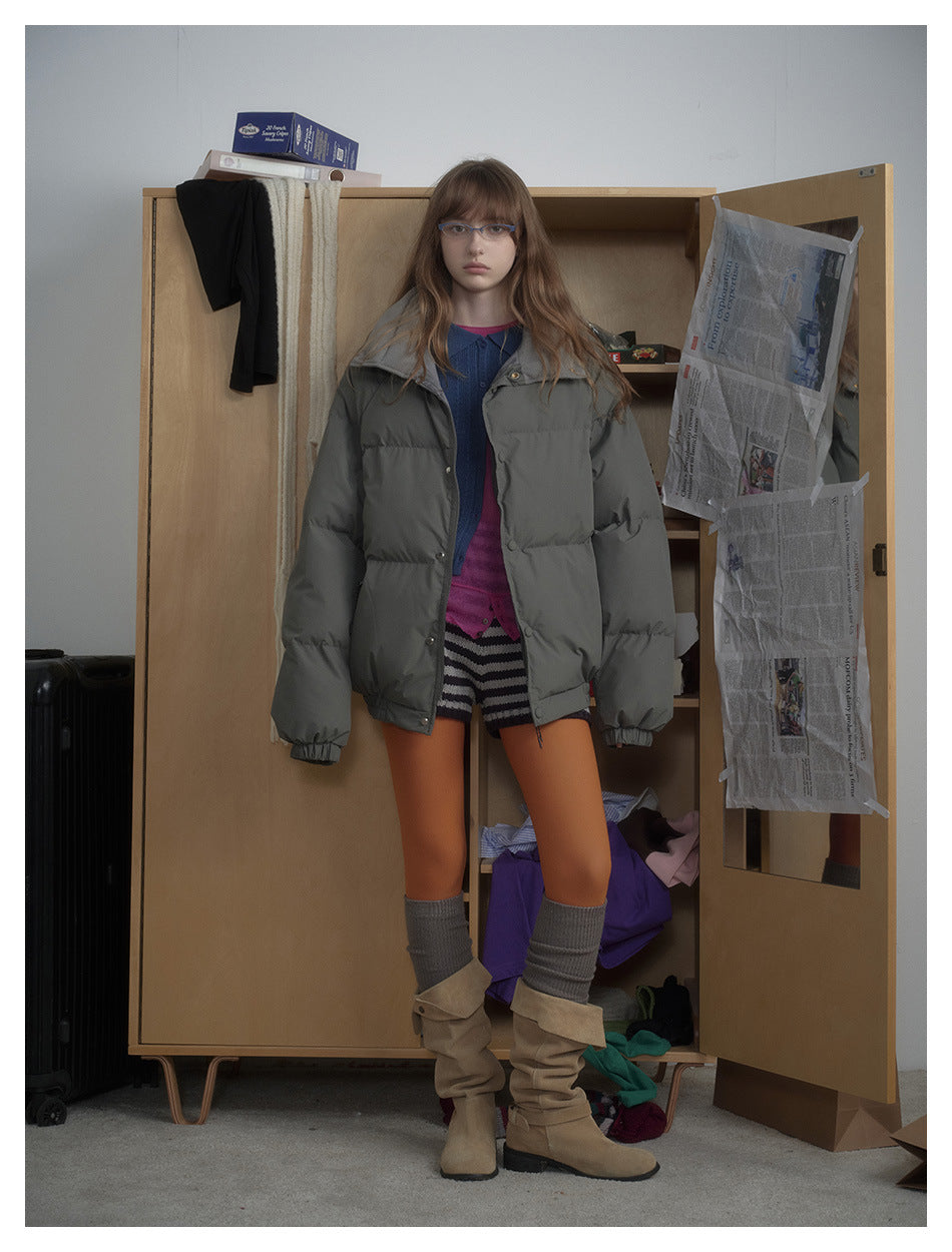 Model wearing an oversized gray puffer jacket open over a blue cardigan, striped shorts and tan boots, standing by a wardrobe