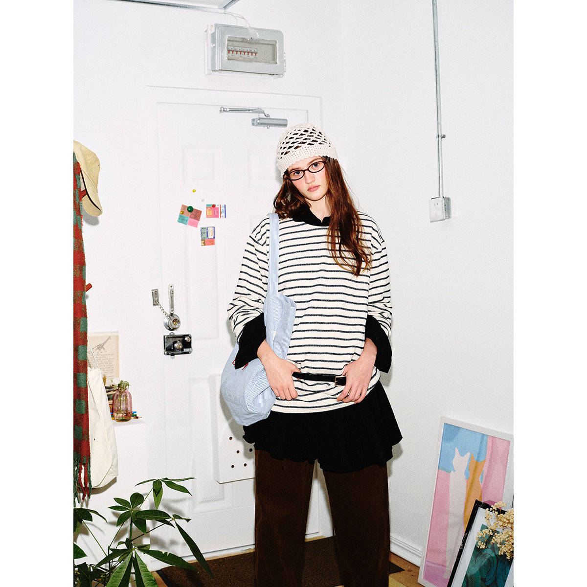 Model wearing an oversized cream shirt with thin dark stripes and black cuffs, layered over a black skirt and brown trousers.